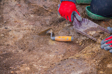 A hand of an archaeologist holding a charcoal-filled container and a trowel lying on the ground.