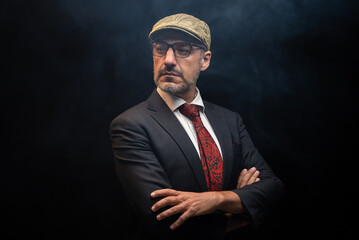 Portrait of detective in elegant black suit and red tie wearing eye glasses and hat standing with crossed arms. isolated on black background.
