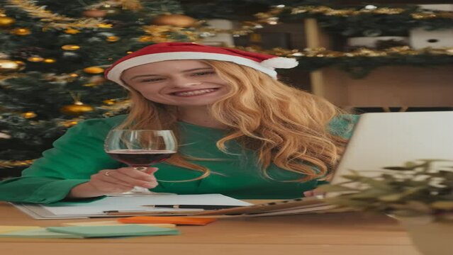 Vertical shot of young blond businesswoman in Santa cap making notes in document, then taking flute of red wine, toasting and making sip while looking at camera against decorated firtree