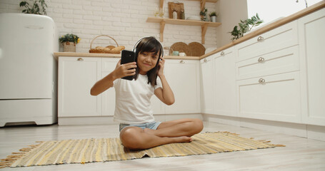 Teen asian girl sitting on floor in kitchen, listening to music in headphones, looking at smartphone and dancing - modern childhood concept 