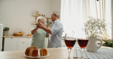Cute senior asian couple are celebrating their anniversary, drinking wine, listening to music and dancing in their kitchen, relaxing after retirement - pension, old couple in love 