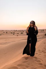 Poster Beautiful mysterious woman in traditional arabic black long dress stands in the desert on sunset © bondvit