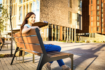 Outdoors lifestyle fashion portrait of brunette woman sitting on the bench. Girl enjoing the sun and resting in urban landscape