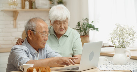 Happy asian senior couple sitting at kitchen table surfing web on laptop together, doing online shopping or watching videos and discussing - happy retirement concept 