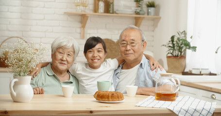 Mature asian couple and their teen granddaughter sitting together at kitchen table, smiling and looking at camera - family ties concept portrait closeup 