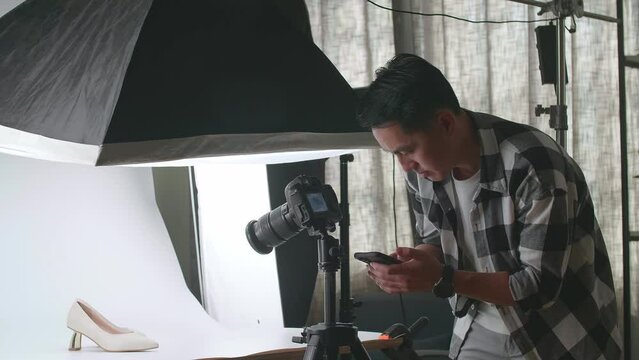 Asian Male Photographer Talking On Smartphone While Taking Photos Of Women'S Shoes In Home Studio 

