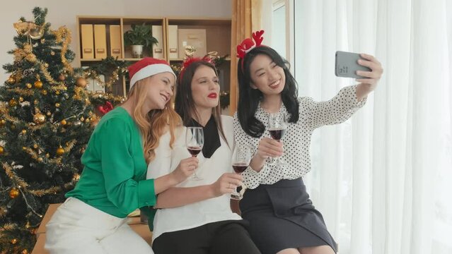 Three happy girls with headbands anf Santa cap toasting for xmas and making video with best wishes on holiday while looking at smartphone screen against decorated firtree