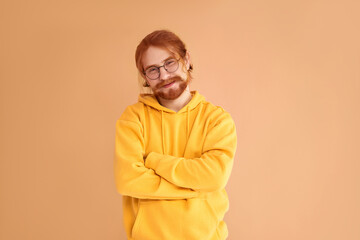 Portrait of a handsome red-haired man with a beard wearing glasses in a yellow hoodie standing with his arms crossed and looking at the camera on a yellow background and smiling.