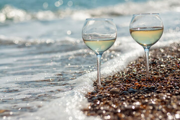 Two glasses of wine on the seashore in the waves and sparkling bokeh from water and pebbles
