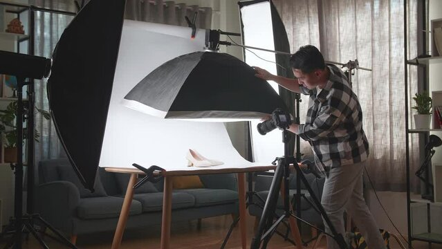 Asian Male Photographer Adjusting Light Equipment To Get Good Illumination On Women'S Shoes While Taking Photos Of The Products In Home Studio 

