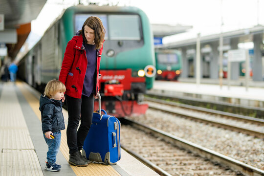 Young mother and her toddler son on a railway station. Mom and little child waiting for a train on a platform. Family with a luggage suitcase ready to travel. Going on vacation with kids.