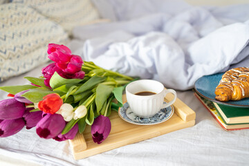 Fototapeta na wymiar Breakfast served with love in bed, coffee, croissant and tulips on a tray on white bed linen at a romantic holiday morning, copy space, selected focus
