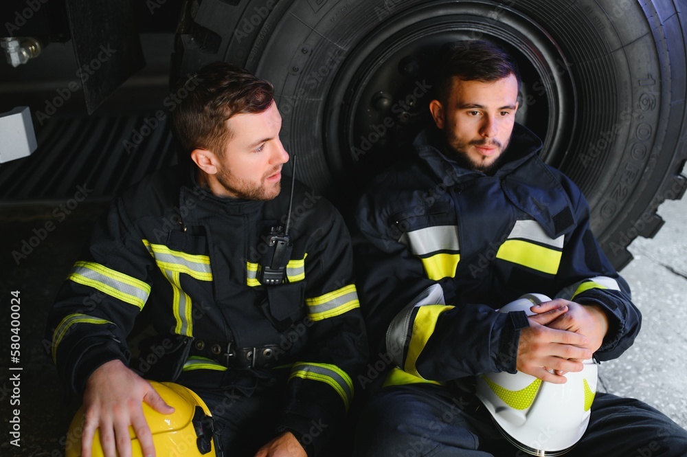 Wall mural two professional firefighters with uniforms and protective helmets posing infront of a firetruck.