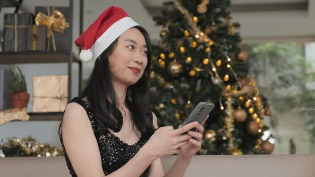 Happy young Asian woman in Santa cap and black dress texting in smartphone while congratulating her friends on Christmas against firtree decorated with sparkling lights