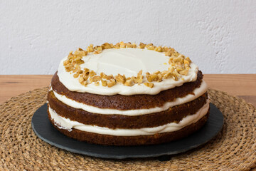 Tarta de zanahoria con glaseado de queso crema y nueces picadas sobre plato de pizarra, mantel individual redondo de mimbre y fondo blanco. 