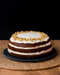 Tarta de zanahoria con glaseado de queso crema y nueces picadas en plato de pizarra, mantel individual redondo de mimbre y fondo negro. Foto vertical, espacio para copiar.