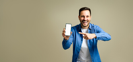 Handsome mid adult man dressed in denim jacket laughing and pointing at smart phone's blank screen...
