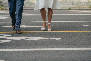 legs of bride in short white dress and sandals and groom in navy blue suit crossing the street in...