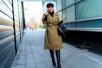 Obraz premium attractive stylish woman talking on the phone and walking down a city street in a trench coat holding purse, elegant style