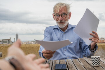 Mature businessman with gray hair and beard in blue shirt, jeans and glasses with paper documents on rooftop terrace with city skylight on background. Copy space