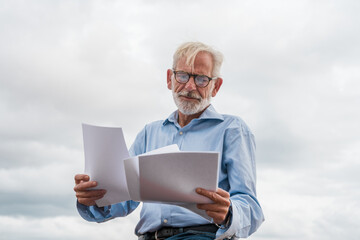 Mature businessman with gray hair and beard in blue shirt, jeans and glasses with paper documents...