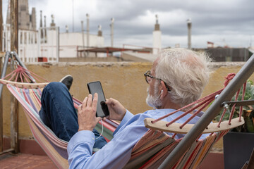 Mature business man with gray hair and beard in blue shirt and jeans laying in hammock with smartphone on terrace