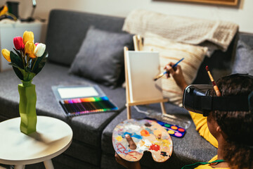 Happy black school girl painting with a paintbrush at home using VR glasses to immerse herself in the metaverse