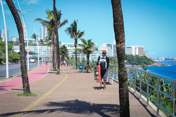 Man with cap and backpack walking on the edge of the beach, next to the bike path, coconut trees and under the blue sky, in the background buildings and apartments
