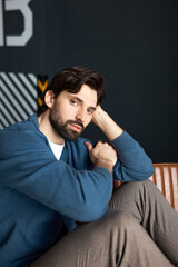 Handsome brutal hipster guy looking at camera with serious confident facial expression, posing in blue cardigan and stylish pants, touching his head, putting elbow on knee. Male fashion