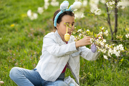 Adorable little girl in bunny ears, blooming tree branch outdoors on a spring day. Kid having fun on Easter egg hunt in the garden