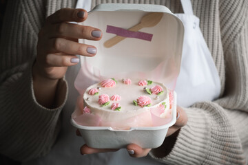 Woman pastry shef wearing white apron and holding small bento cake decorated with cream cheese pink flowers. Woman hands hold birthday cake in a white gift box. Trendy Korean style cake