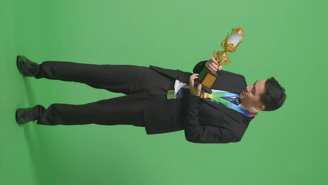 Full Body Of Asian Business Man In A Suit And Tie With A Gold Medal Looking At A Gold Trophy In His Hands And Saying Wow Being Happy Winning As The First Winner On Green Screen Background In The Studi