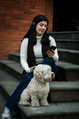 happy woman with her dog listening to music in the city. Happy girl with her dog looking at the cell phone. The dog looks into the distance. A girl sitting with her dog on the stairs