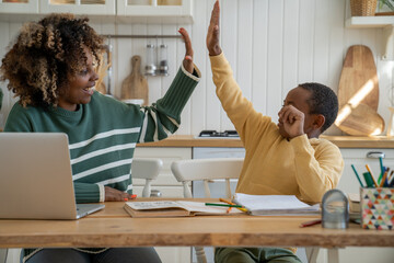 Homeschooling and freelancing. Happy African American child son giving high five to mother while doing homework or studying together at home, sitting at kitchen table with books and laptop