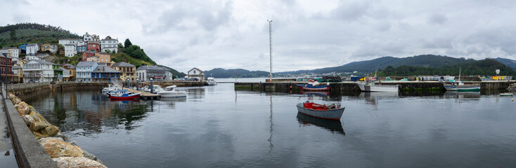 Fototapeta na wymiar Panoramic view of the village of O Porto do Barqueiro, Galicia, with boats moored at the pier, reflected in the blue water of sea, mountain in the background with houses traveling in summer of 2021