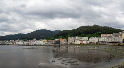 Obraz premium Vistas panorámicas de la ría de Viveiro en Lugo, con marea baja y arena mojada reflejando los edificios de la orilla, montañas verdes y cielo de verano nuboso en España 2021