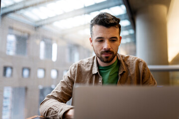 Portrait of pensive, handsome Latin student using laptop computer, Freelancer working in modern office. Attractive copywriter sitting at workplace. Successful business - obrazy, fototapety, plakaty