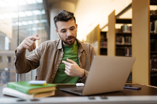 Handsome man using laptop, explaining something during video call. Middle aged student in wireless earphones has an online lesson, recording a video. Freelancer working online sitting at workplace