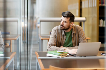 Pensive, attractive man using laptop working online, sitting at the workplace. Hipster guy in...