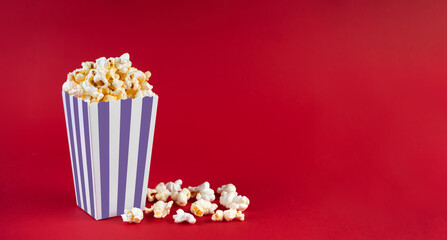 Purple white striped carton bucket with tasty cheese popcorn, isolated on red background. Box with scattering of popcorn grains. Fast food, movies, cinema and entertainment concept.