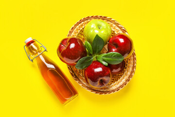 Glass bottle of fresh apple cider vinegar and basket with fruits on yellow background
