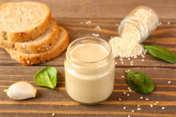 Jar with tasty tahini, sesame seeds, bread and spinach on wooden table