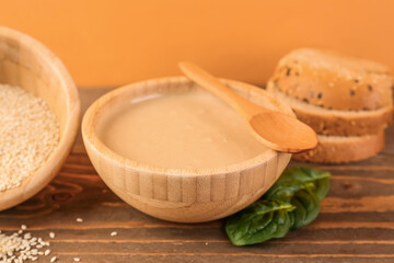 Bowls with tasty tahini, sesame seeds, bread and spinach on wooden table near color wall, closeup