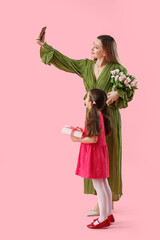 Cute little girl with her mother and gifts taking selfie on pink background
