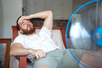 Young bearded man using electric fan at home, sitting on couch cooling off during hot weather,...