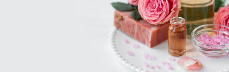 Aromatherapy. Pure organic essential rose oil concept. Elixir with plant based floral herbal ingredients. Pink flowers extract. Spa atmosphere with candle, towel. White background. Banner copy space