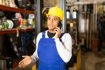 Portrait of hispanic worker of building materials store in blue coverall and hard hat talking on...