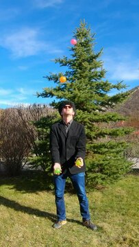 Vertical slow motion : man juggling with four balls of colors (orange, green, pink, yellow) in front of a pine tree.