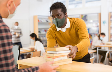 Portrait of young adult man wearing face mask for viral protection returning books to librarian at...