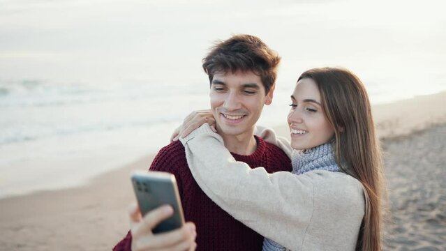 Video of beautiful young couple making photos with mobile phone in a cold winter on the beach.